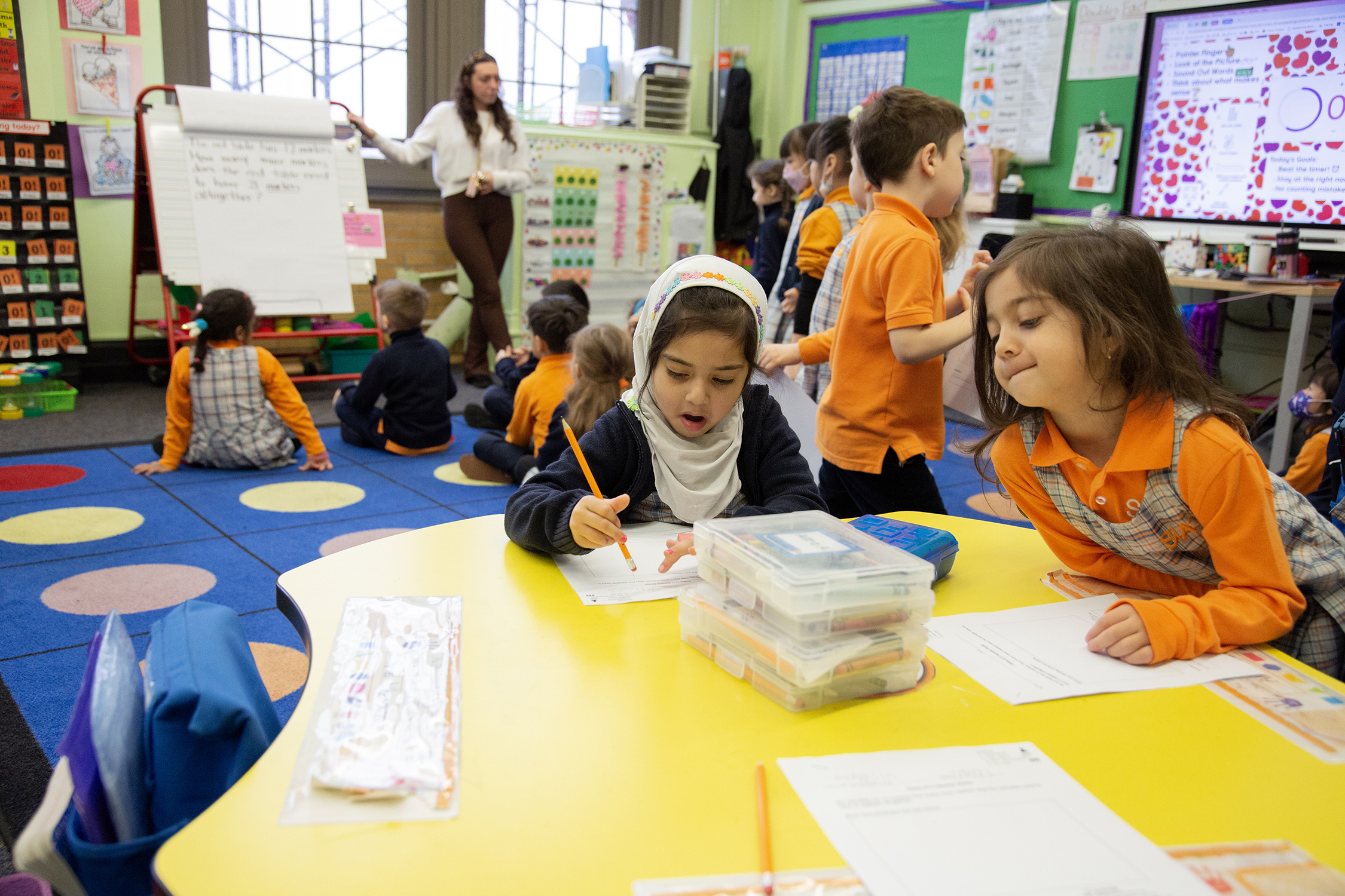 Kindergarteners prepare worksheets while their teacher waits for everyone to come to the rug