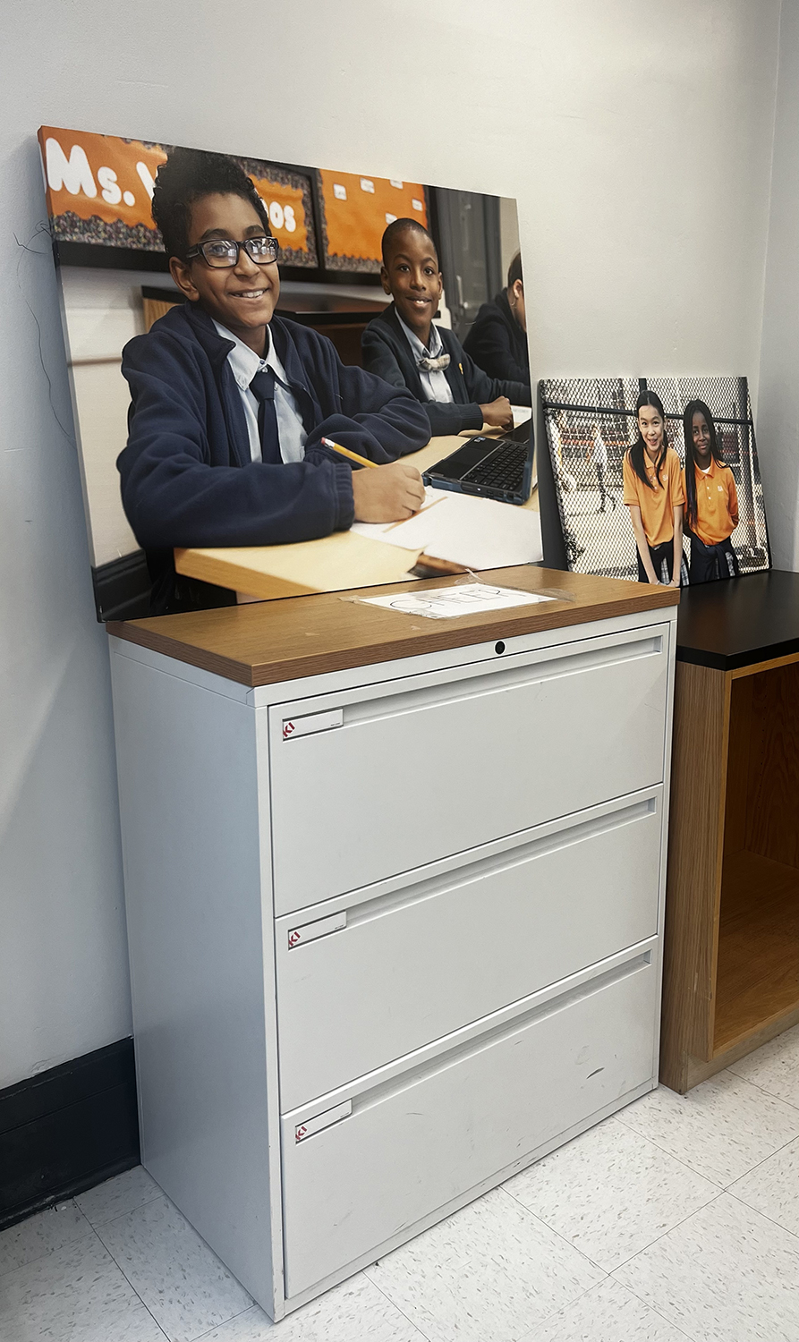 two canvas displayed on filing cabinets show four children smiling in classroom and next to chainlink fence